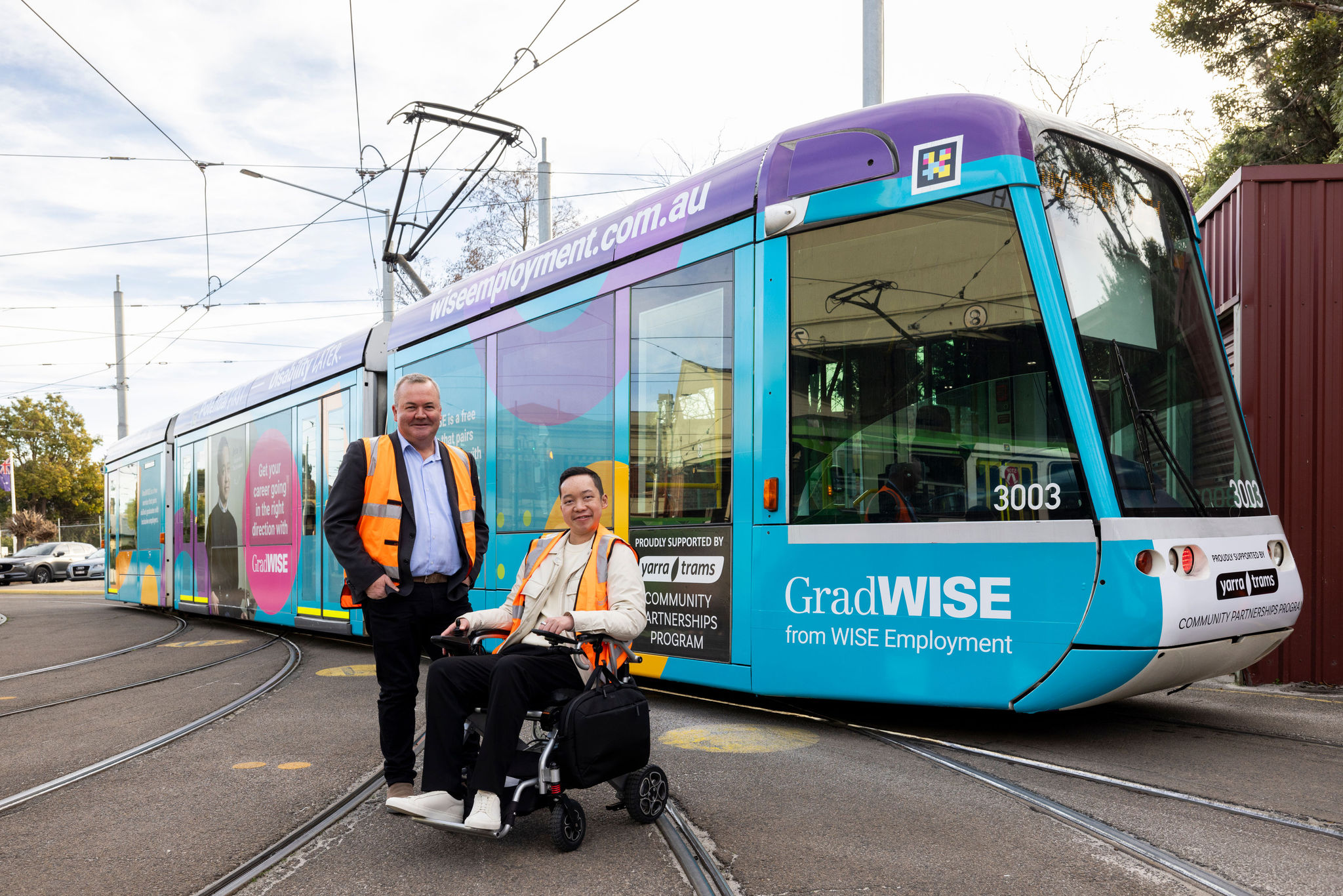 Two men in high vis vests pose infront of a C1 tram, one man is standing next to the other who is in a motorised wheelchair. The words 'GradWISE from WISE Employment' are printed on the front of the tram.