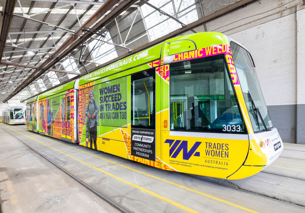 Photo of a C1 tram in a maintenance shed, wrapped in a yellow, green and pink design from Tradeswomen Australia, including the wording 'Women Succeed in Trades and you can too'