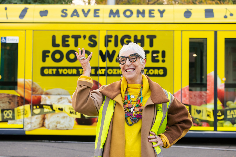 Photo: Ronni Kahn AO, OzHarvest CEO and Founder poses in front of a vibrant yellow tram, which has images of food and the text 'Save Money' and 'Let's Fight Food Waste' in black lettering.