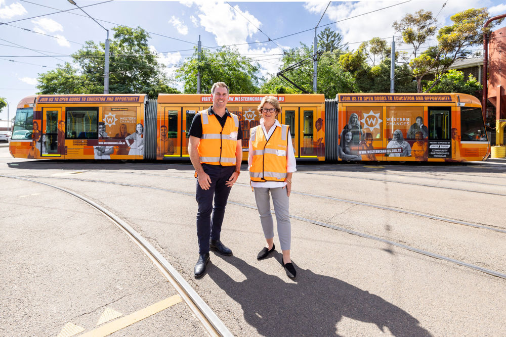 Audio: Photo of Terry Bracks AM and Zac Lewis posing in front of a colourful tram showcasing the faces of scholarship recipients, with the text “Join us to open doors to education and opportunity”