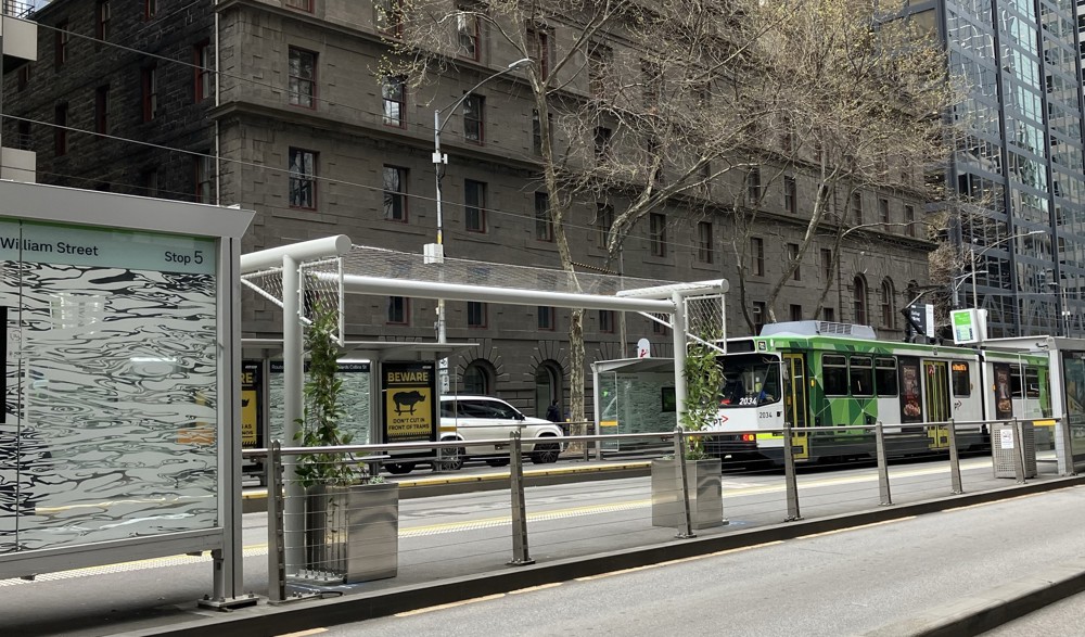 Photo of a tram platform, showing a standard tram shelter alongside an 'urban arbour' - a white shelter structure with green vines growing up the sides.