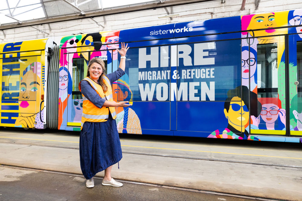 Photo: A SisterWorks employee in a high visibility vest poses in front of a vibrantly coloured tram with the text 'Hire Migrant & Refugee Women' printed.