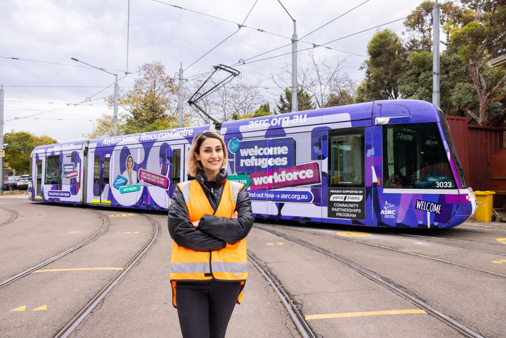 Woman in an orange vest stands in front of a purple tram with large text spelling out 'WELCOME' printed on it