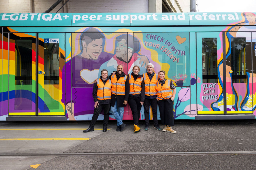 Five Switchboard representatives in high visibility vests pose in front of the Switchboard C class tram with the words 'LGBTIQA+ peer support and referral', 'check in with your friends' and 'play with pride'
